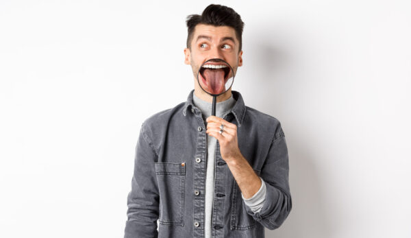 Handsome positive guy showing white perfect teeth and tongue with magnifying glass, looking left at logo, standing against white background.