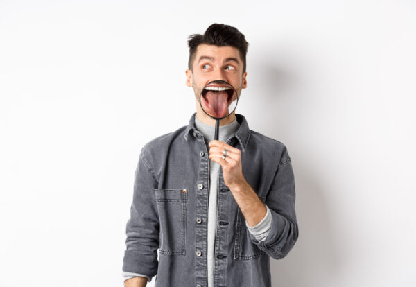 Handsome positive guy showing white perfect teeth and tongue with magnifying glass, looking left at logo, standing against white background.
